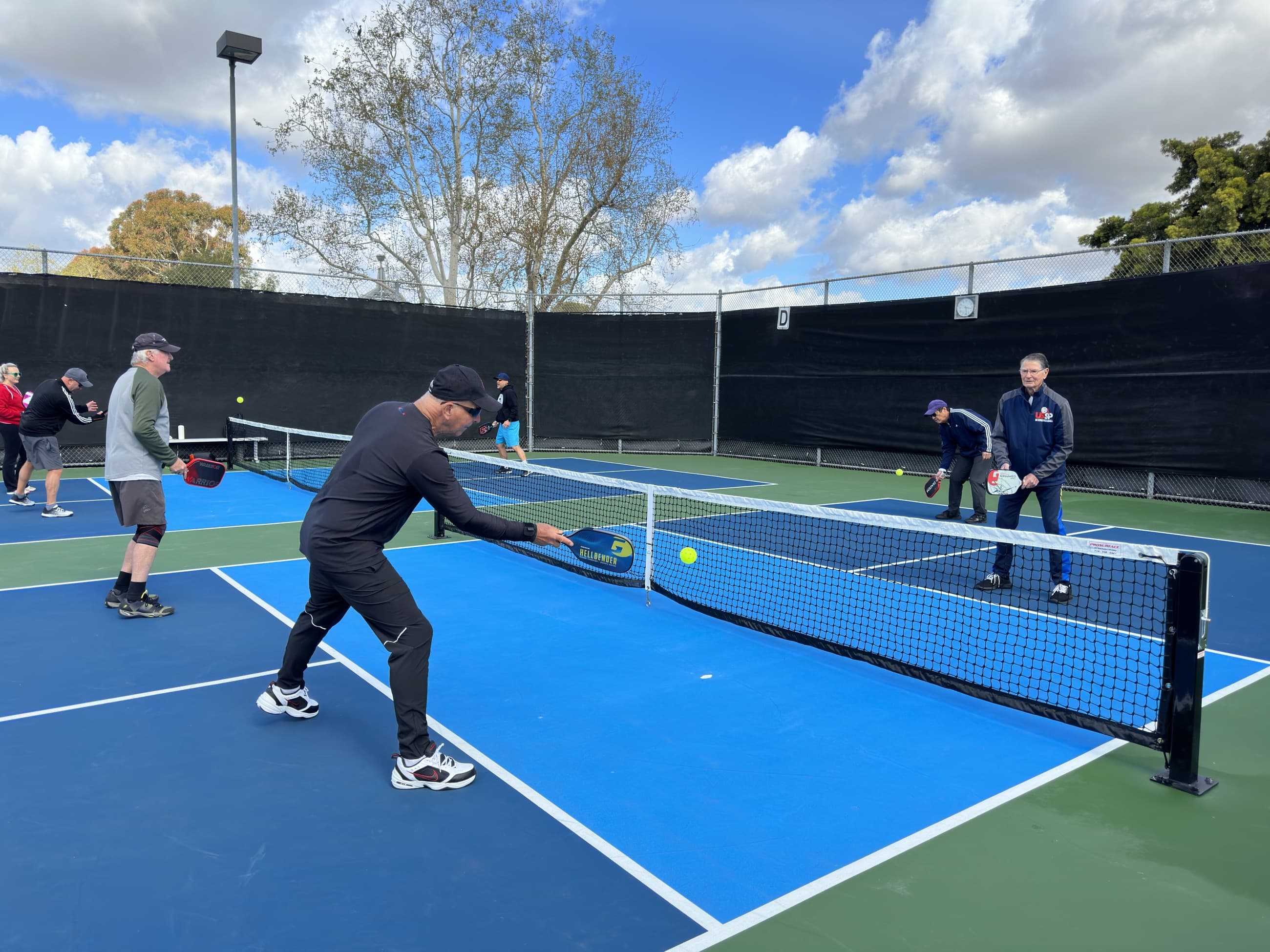 Players rally across a bright blue-and-green outdoor court under open sky.